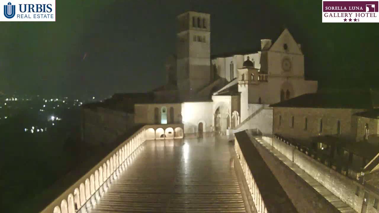 A large, illuminated stone building complex sits atop a hill at night under a dark, possibly rainy sky, overlooking a city with many lights in the distance.