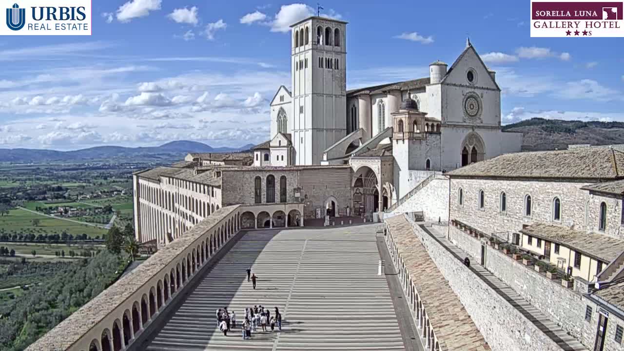 A large, light-colored stone basilica and attached buildings are seen from above on a sunny day, with a wide plaza stretching out from its entrance and a hilly landscape visible in the distance.