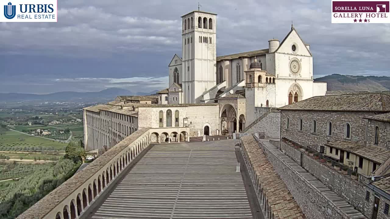 A large, light-colored basilica and adjoining buildings overlook a wide, paved plaza and a hilly landscape under a cloudy sky.