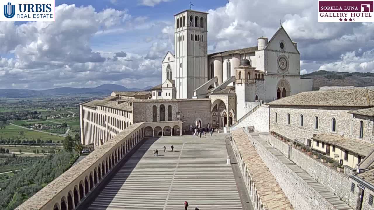 A large, light-colored stone basilica and adjoining buildings are shown on a sunny day with partly cloudy skies, overlooking a sprawling landscape.
