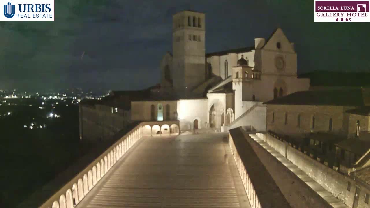 A nighttime view of a large, illuminated stone building complex, possibly a church or monastery, with a long, sloped walkway leading to it, under a partly cloudy sky.