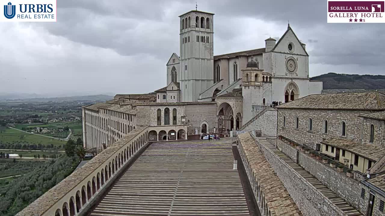 A large stone basilica and adjacent buildings are viewed from above on an overcast day, with a wide paved area and a surrounding landscape visible in the distance.