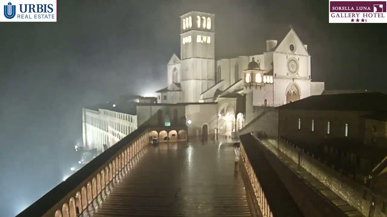 A large, illuminated basilica is seen at night under foggy conditions, viewed from a high vantage point across a wet, paved area between two long, arched walkways.