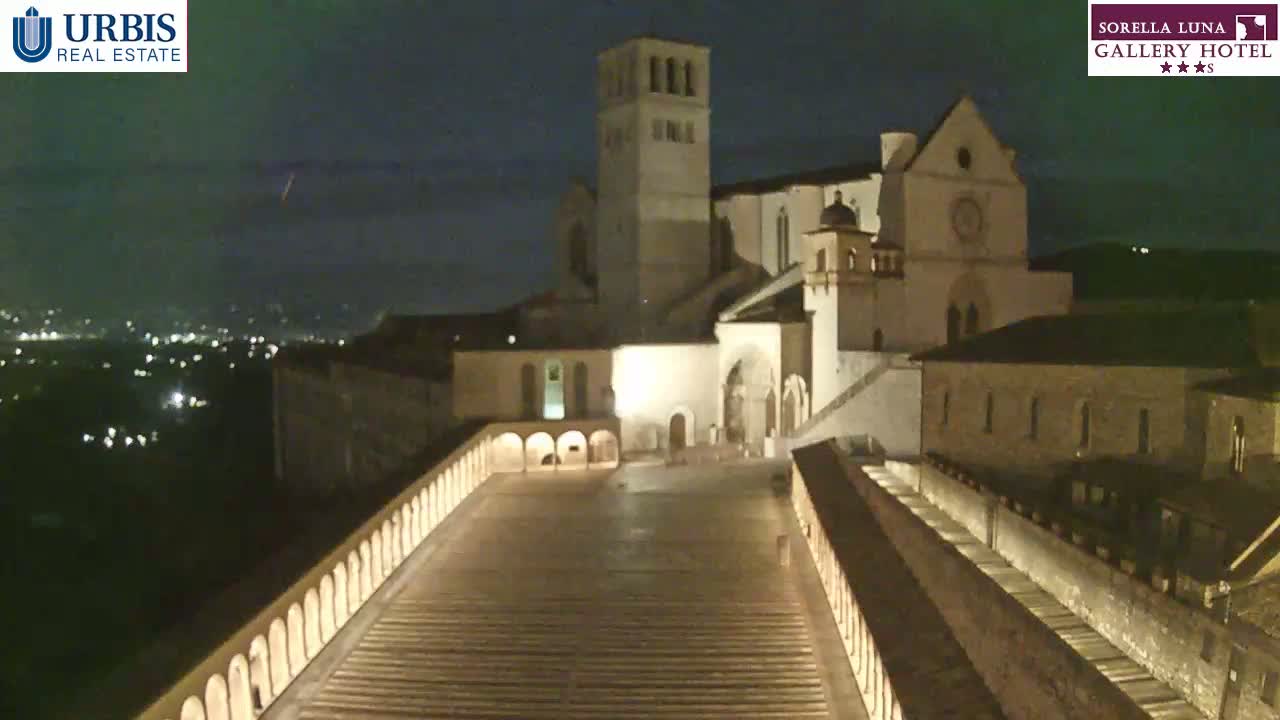 A large, illuminated stone basilica sits atop a hill at night, overlooking a city with distant lights, under a dark sky.