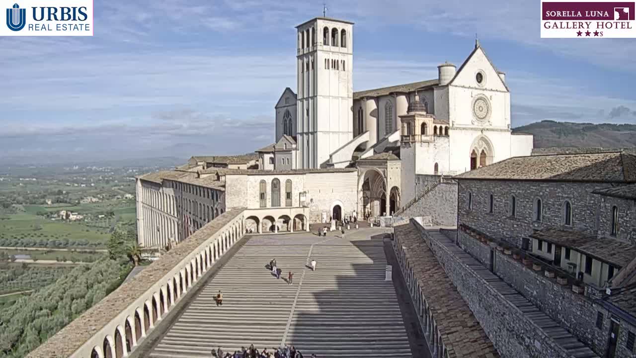 A large, light-colored basilica with a tall bell tower stands overlooking a wide, descending staircase and a sprawling landscape under a mostly clear sky.