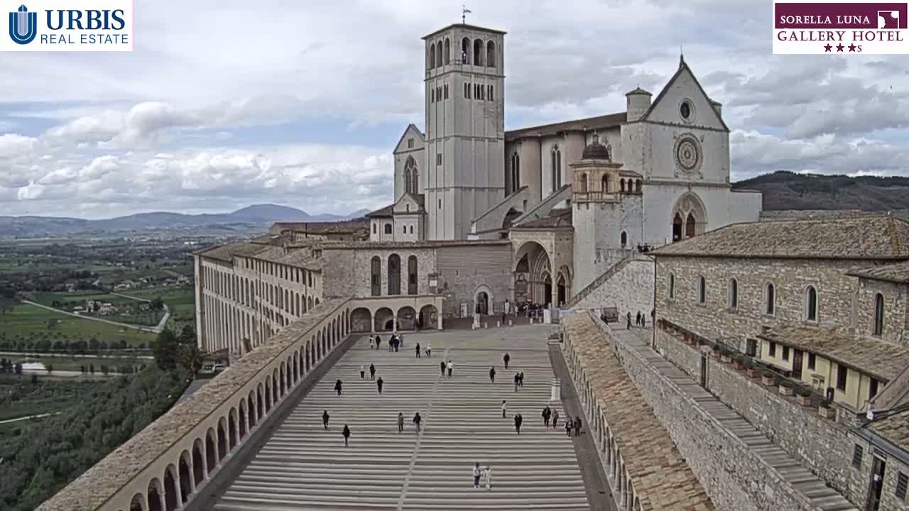 A large stone basilica and adjoining buildings are viewed from above on a partly cloudy day, with a wide plaza and long stairways leading to the main entrance.