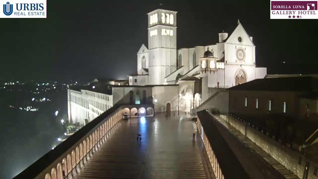 A large, illuminated basilica sits on a hill at night, overlooking a wet plaza surrounded by colonnades, under a dark sky.