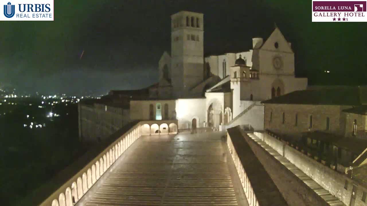 A nighttime view of a large, illuminated stone building complex, possibly a church or monastery, overlooking a city with lights visible in the distance under a dark, clear sky.