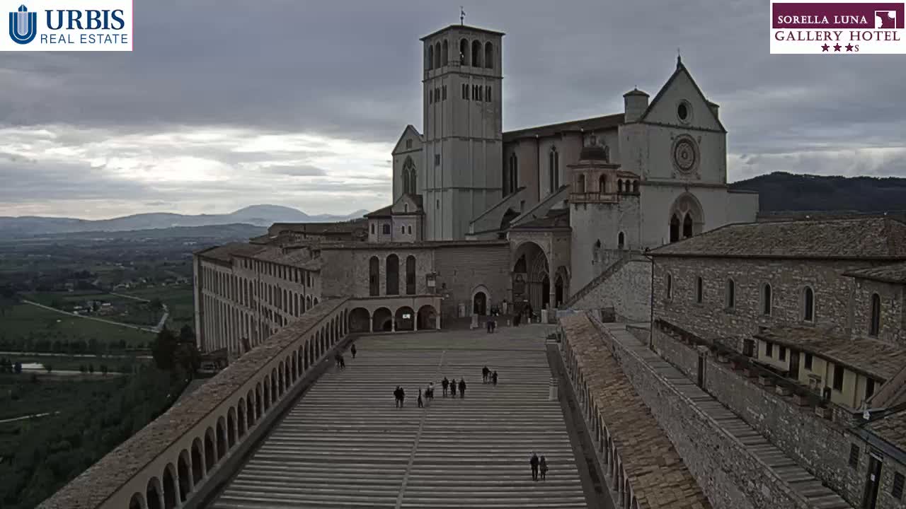 A large stone basilica and adjacent buildings are shown from an elevated viewpoint on an overcast day, with a wide staircase leading to the basilica's entrance and a valley visible in the background.
