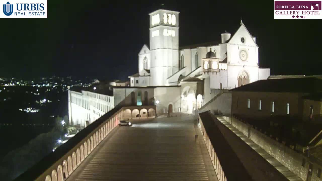 A large, illuminated basilica stands at night overlooking a city, viewed from a high vantage point along a long, arched walkway.