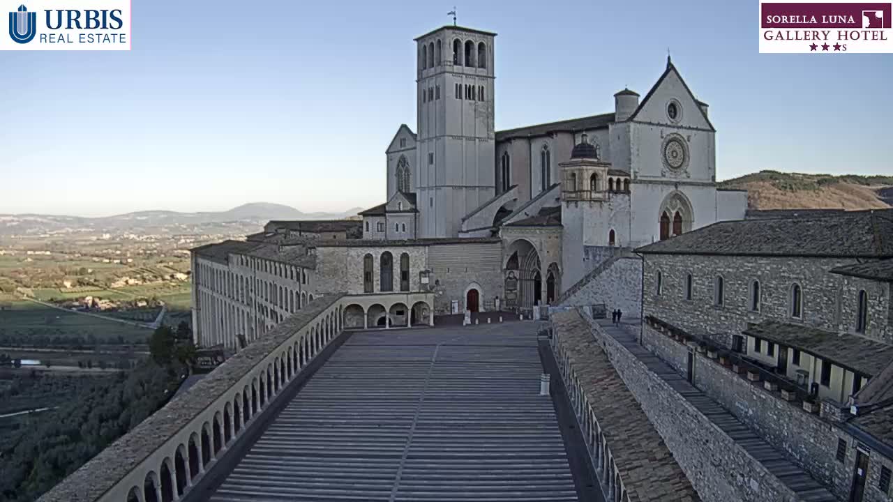 A large stone basilica and surrounding buildings are viewed from above on a clear, sunny day, overlooking a valley in the distance.