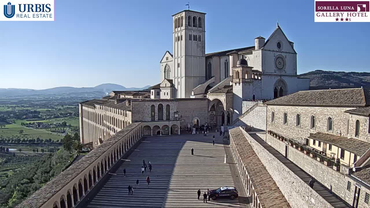 A large stone basilica with a tall bell tower overlooks a paved plaza and a sprawling landscape under a clear blue sky.