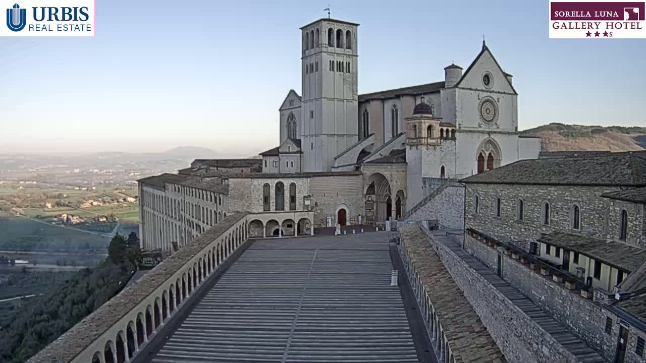 A large stone basilica and adjoining buildings overlook a valley under a clear, bright sky.