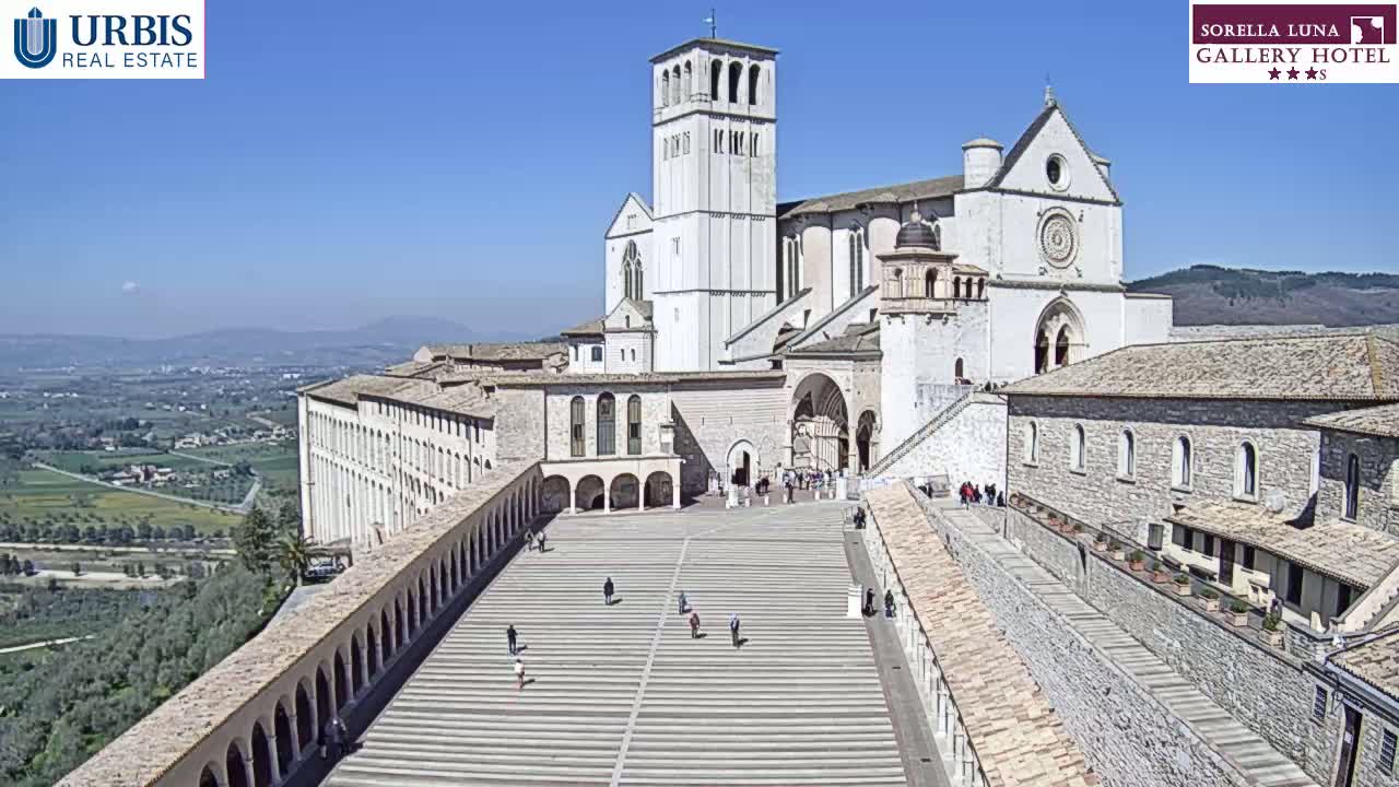 A large, light-colored stone basilica and adjoining buildings are seen on a sunny day, with a spacious plaza in the foreground and a panoramic view of a valley in the background.