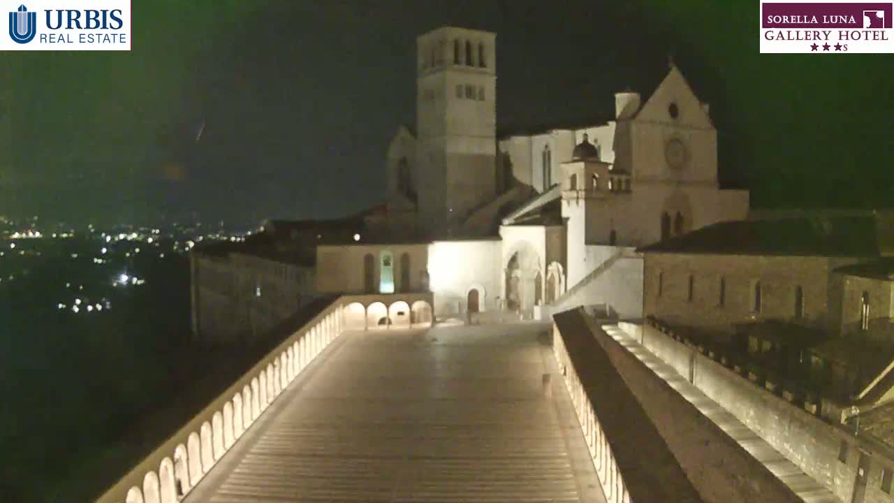 A large, illuminated basilica sits atop a hill at night, overlooked by a long, sloped walkway flanked by low walls, with a distant city glowing faintly in the dark.