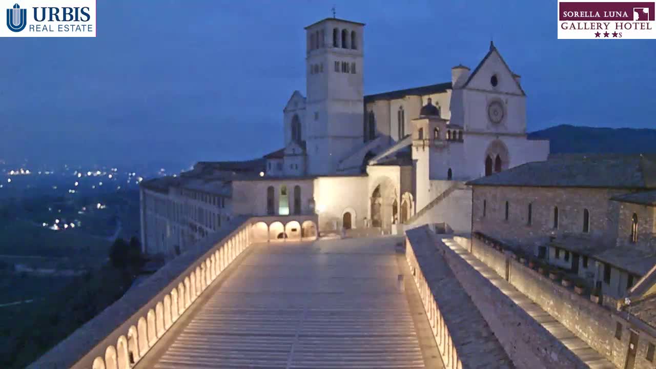 A large, illuminated basilica complex sits on a hilltop at dusk, overlooking a city with lights visible in the distance.