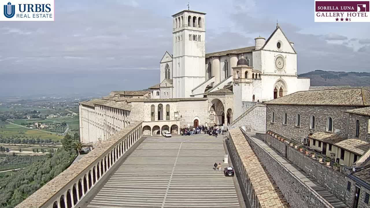 A large stone basilica and adjoining buildings are viewed from above on a partly cloudy day, with a wide plaza and surrounding landscape visible in the distance.