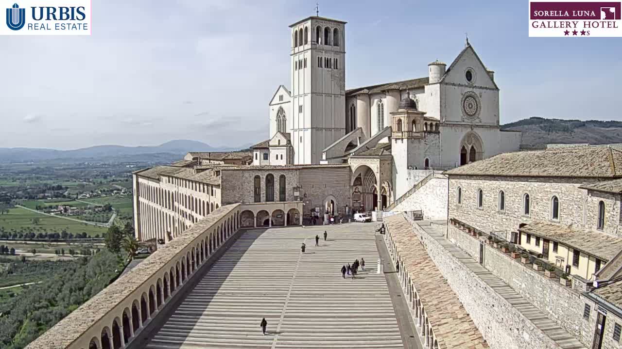 A large, light-colored stone basilica sits atop a hill overlooking a sprawling valley on a sunny day, with a wide plaza leading up to its entrance.