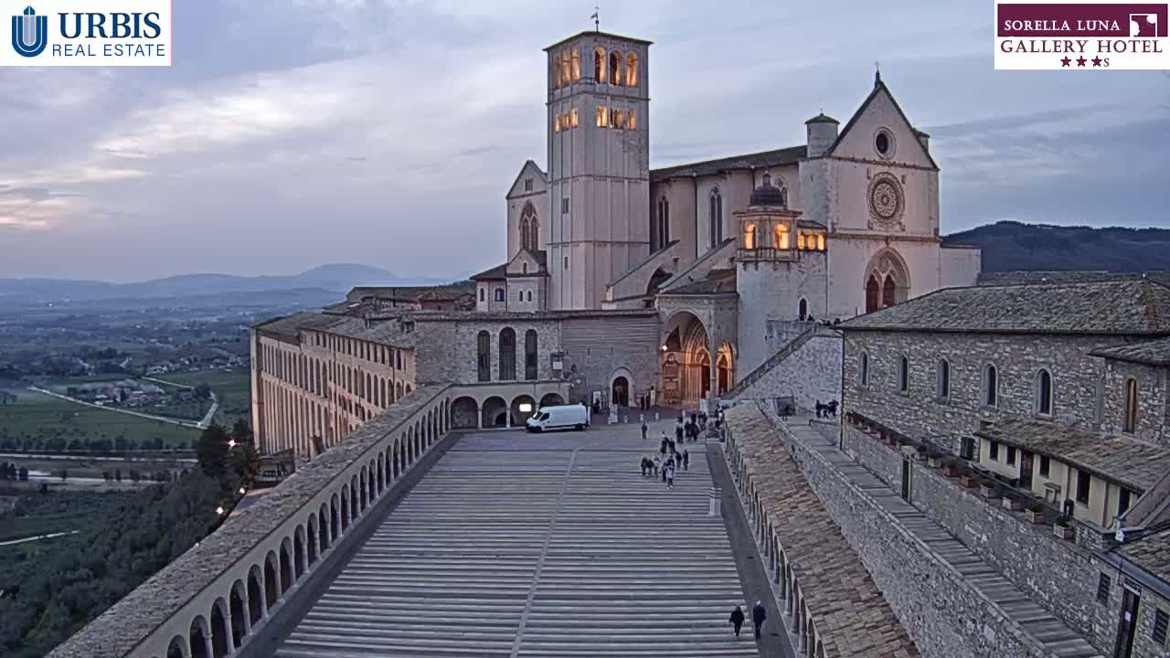 A large stone basilica and adjoining buildings are situated on a hilltop overlooking a valley at dusk.