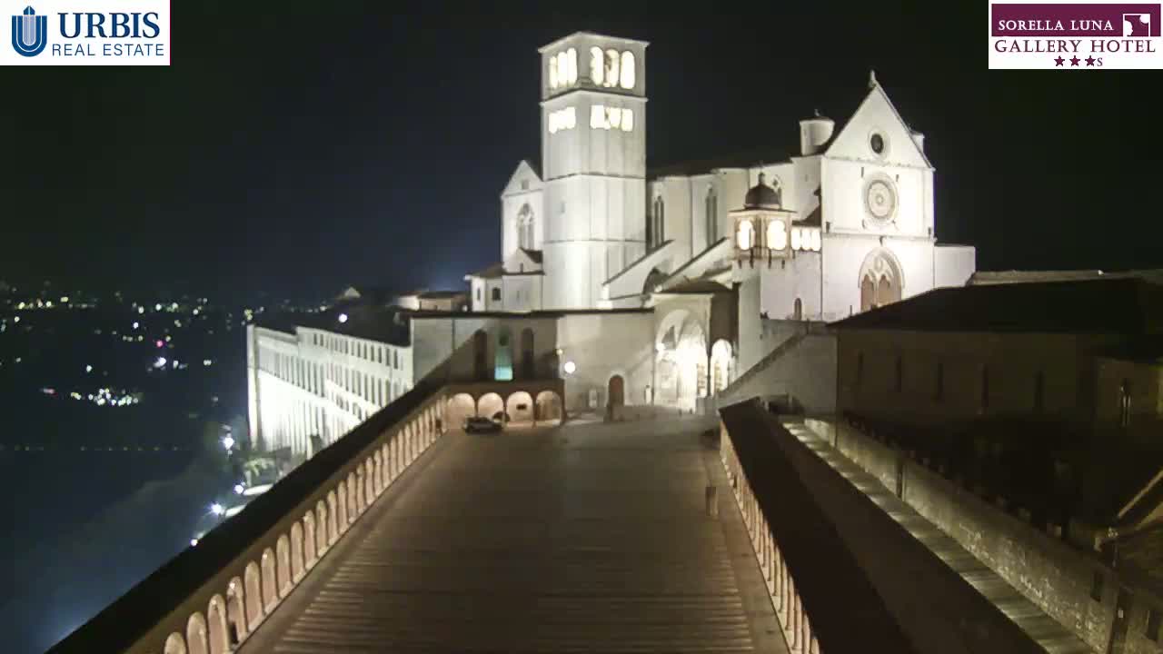 A large, illuminated basilica stands at night, overlooking a city with a long, stone walkway leading to it.