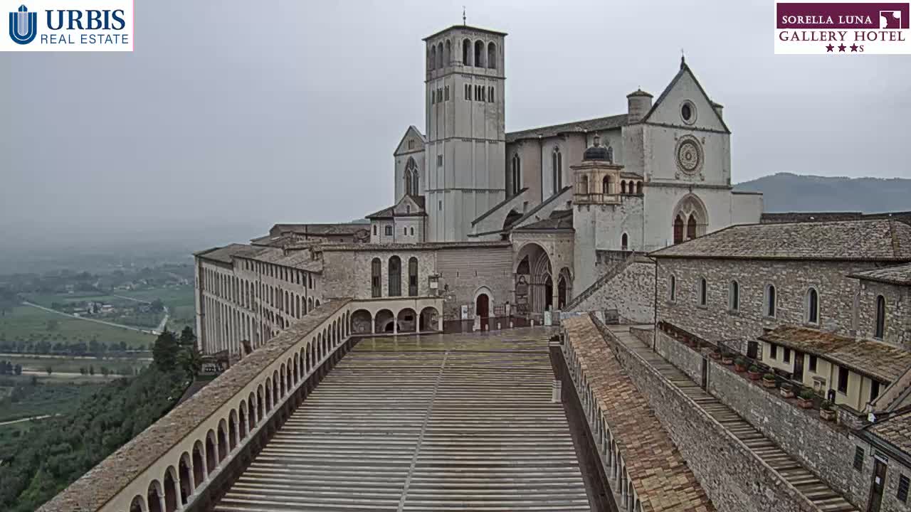 A large stone basilica and adjoining buildings overlook a wide, paved plaza on an overcast day.