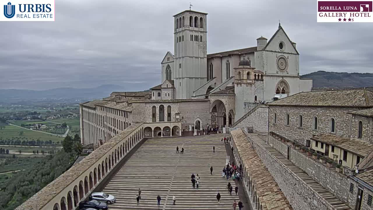 A large stone basilica and adjoining buildings are seen from an elevated viewpoint on an overcast day, with a wide staircase and walkway leading down to a plaza filled with people.