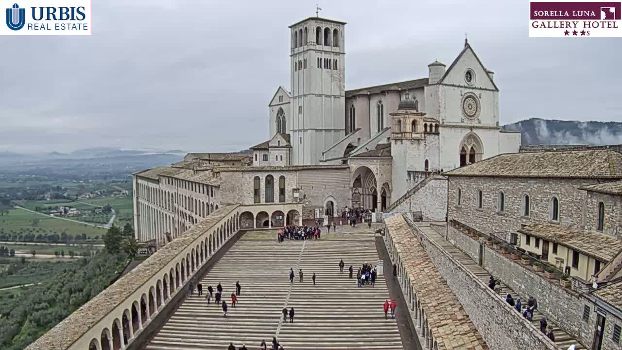 A large stone basilica and adjoining buildings are seen from above on an overcast day, with people walking on a long set of stairs leading to the main entrance.