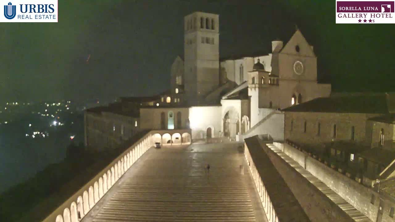 At night, a large, illuminated stone complex with a tall tower and a wide, sloped plaza is seen from an elevated viewpoint.
