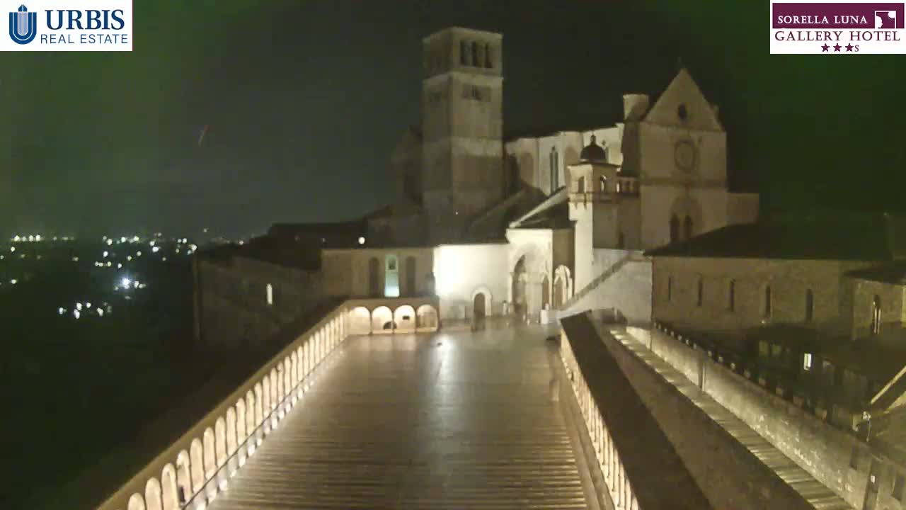 A nighttime view of a large stone building complex, possibly a church or monastery, with a long, elevated walkway leading to it, under a clear, dark sky.
