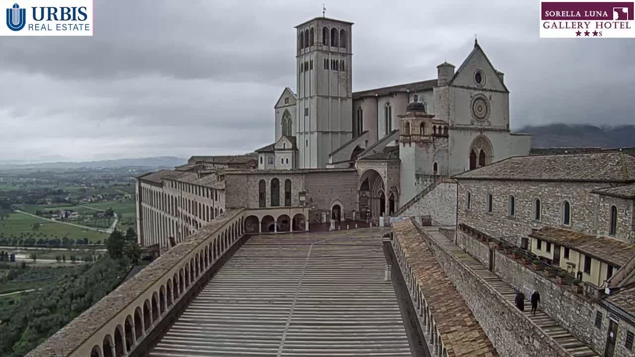 A large stone basilica complex is shown on an overcast day, with a long paved courtyard extending from the main building towards the viewer, surrounded by arched walkways and additional structures.