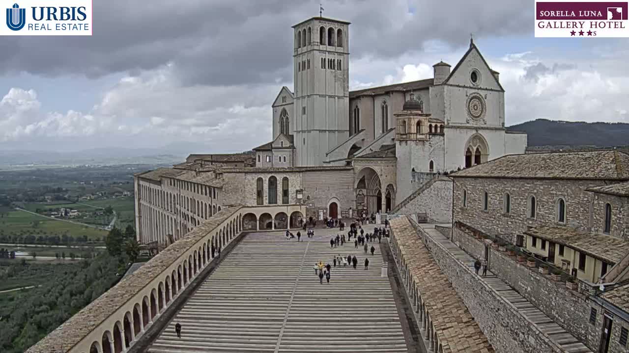 A large stone basilica and adjoining buildings are seen from above on a cloudy day, with many people walking in the plaza between them.