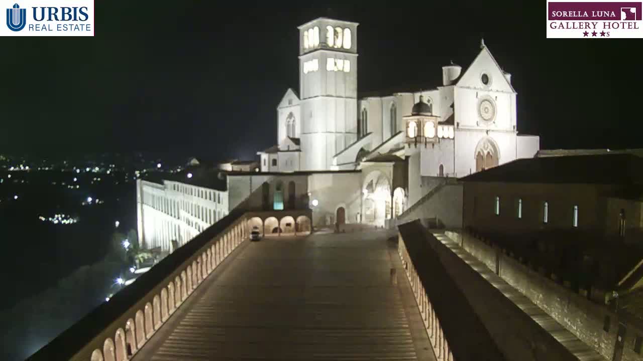 A large, illuminated basilica stands at night, overlooking a city with a long, arched walkway leading to it.