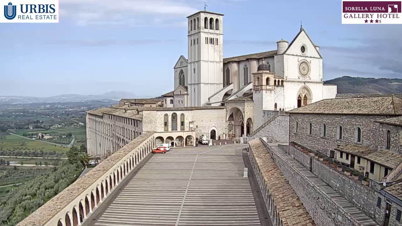 A large, light-colored basilica with a tall bell tower sits atop a hill overlooking a sprawling valley under a mostly clear sky.