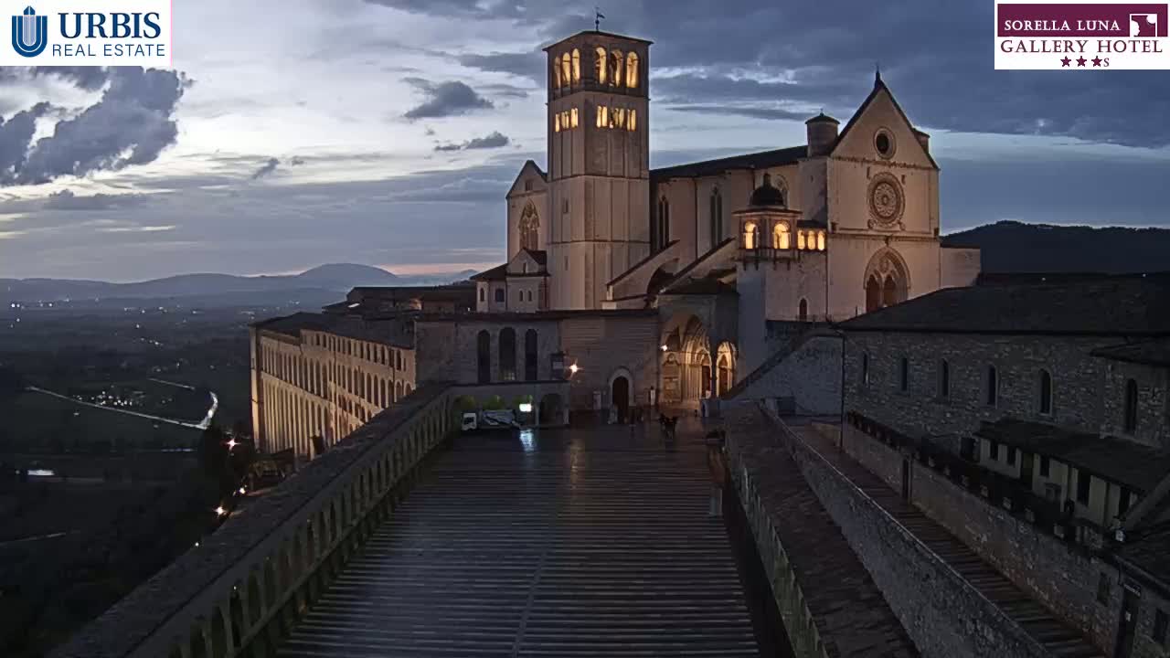 A large, illuminated basilica stands on a hilltop at dusk, overlooking a valley under a cloudy sky.