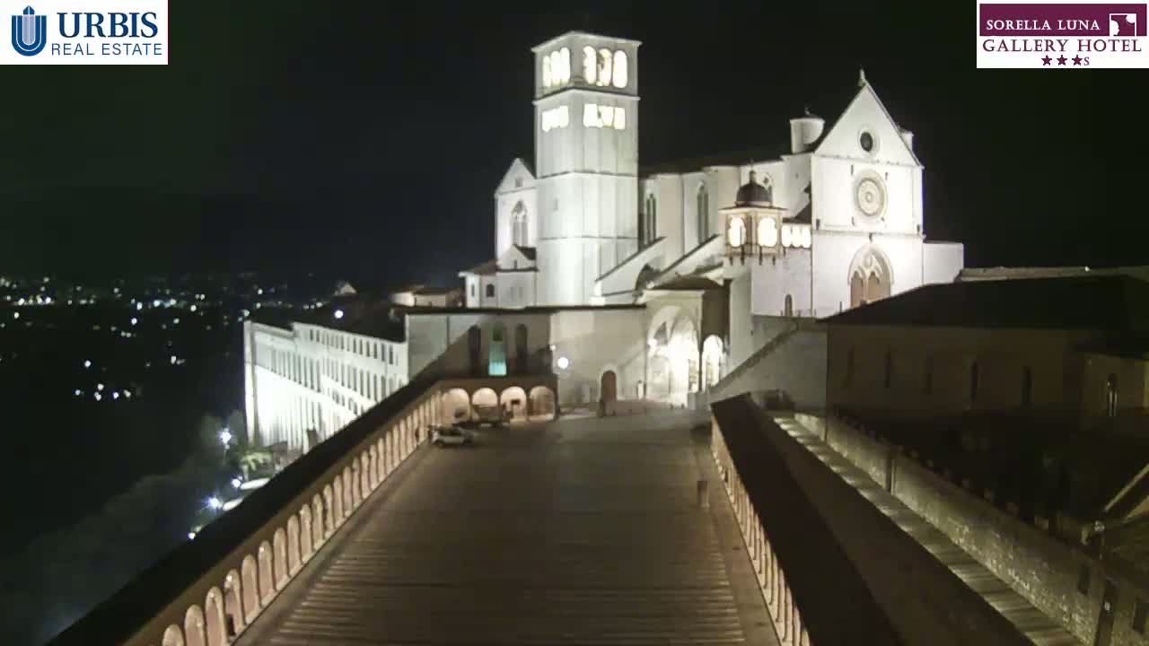 At night, a large, illuminated basilica sits atop a hill overlooking a city, with long, arched walkways extending from the basilica down the hillside.