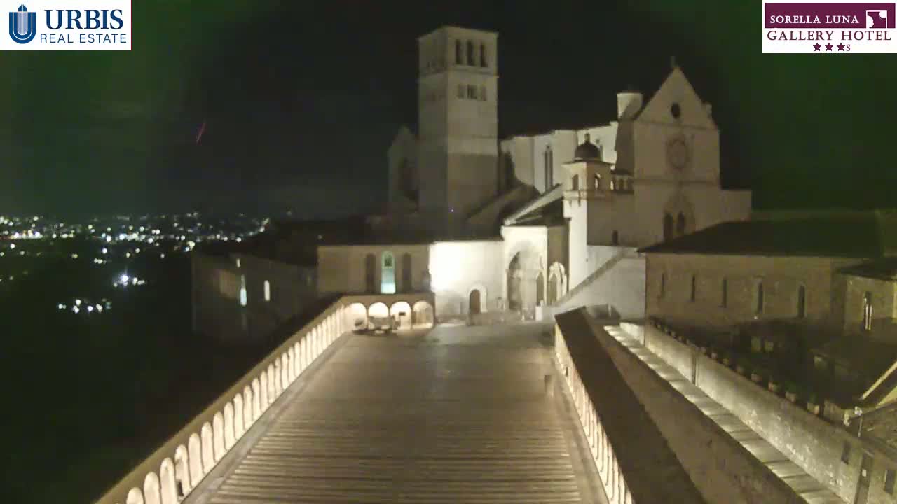 A nighttime view of a large stone building complex, illuminated, with a long, descending walkway in the foreground and a city spread out in the distance under a dark sky.