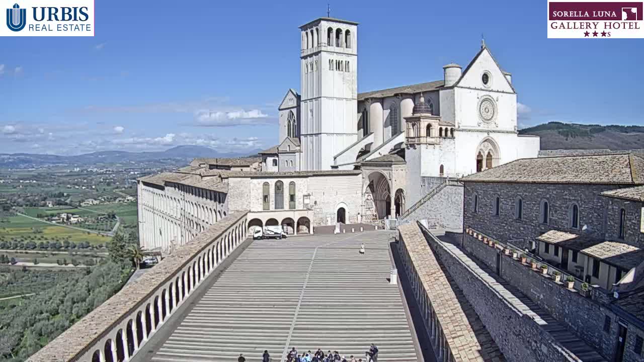 A large, light-colored basilica and adjoining buildings are viewed from above on a sunny day, with a sprawling, paved plaza and distant hills visible in the background.