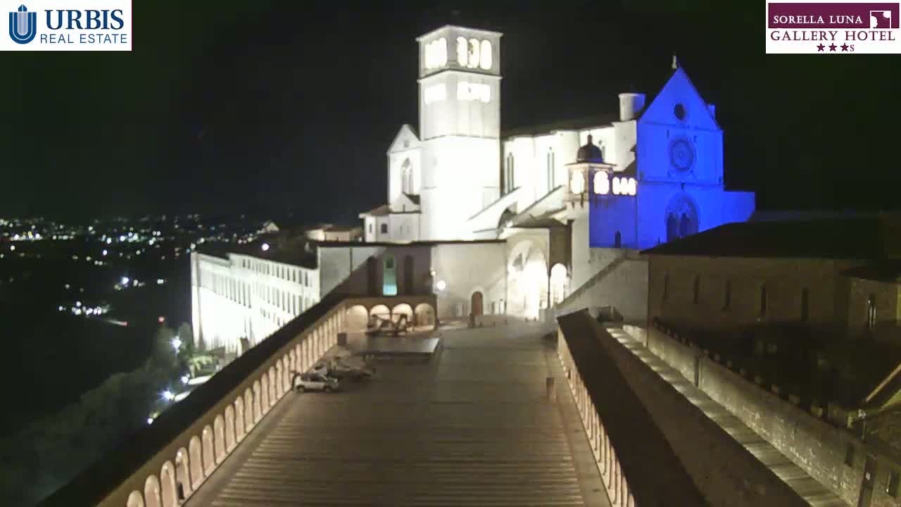 A large, illuminated basilica is seen at night, with a city sprawling in the distance under a clear, dark sky.