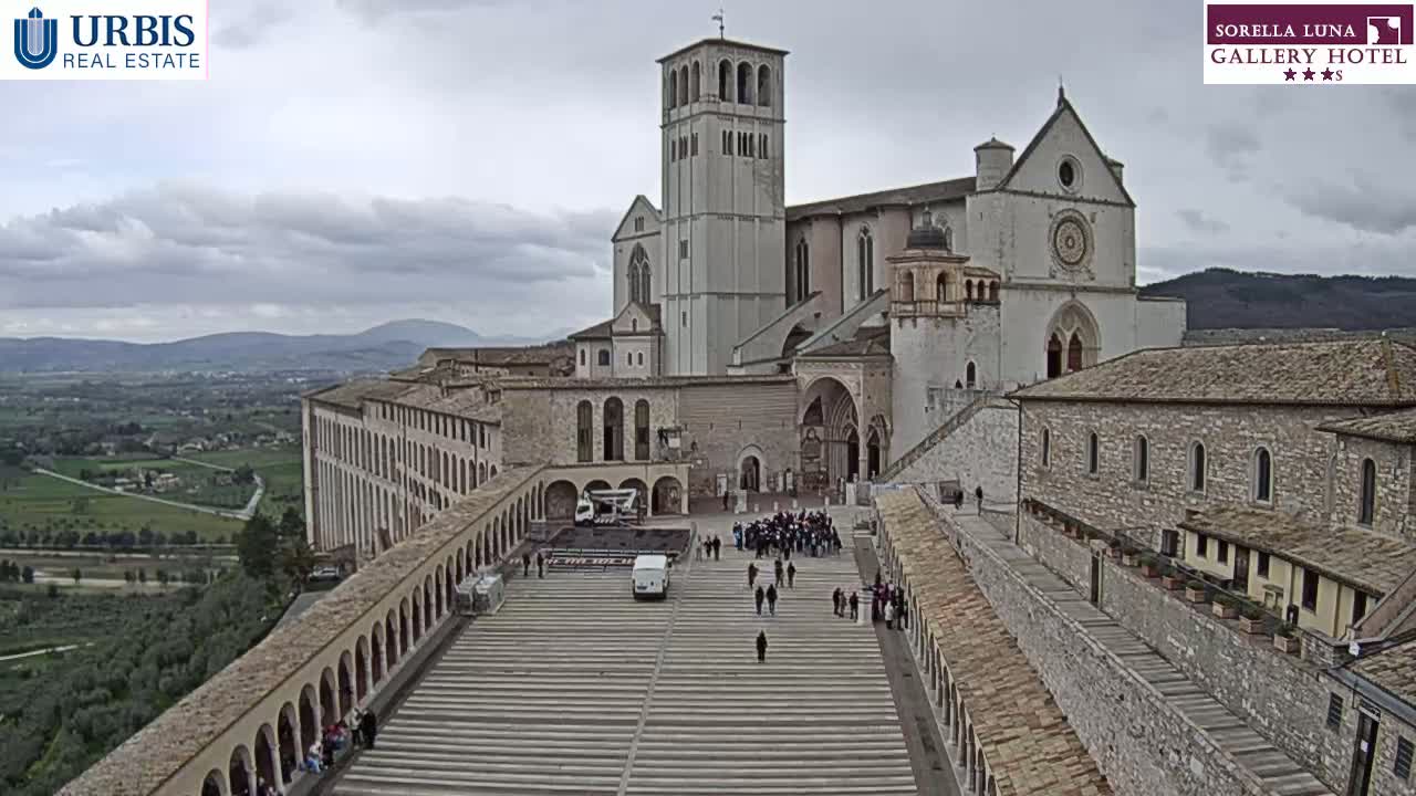 A large stone basilica and surrounding buildings are viewed from above on an overcast day, with a crowd of people gathered in the plaza below.