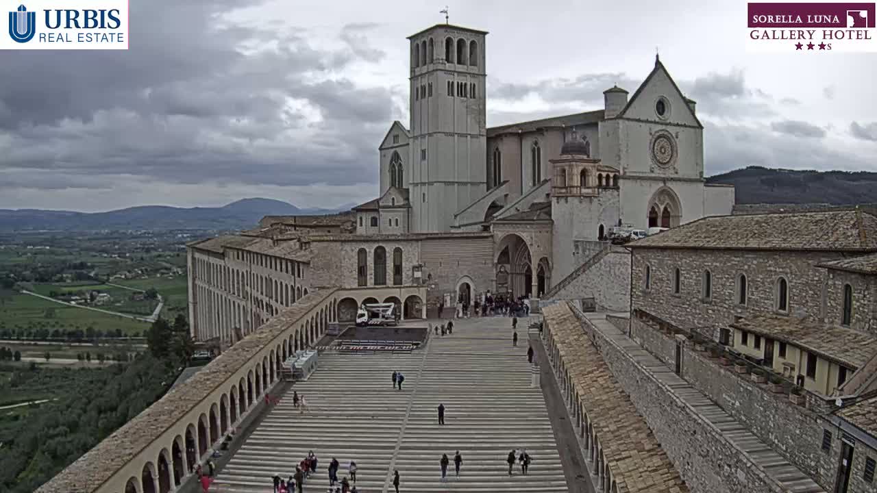 A large stone basilica and adjacent buildings are shown on an overcast day, overlooking a wide, paved area with people walking, and a valley visible in the background.