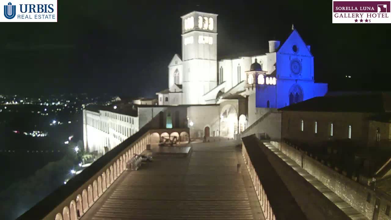 A large, illuminated basilica is viewed from above at night, with a city visible in the distance under a clear, dark sky.