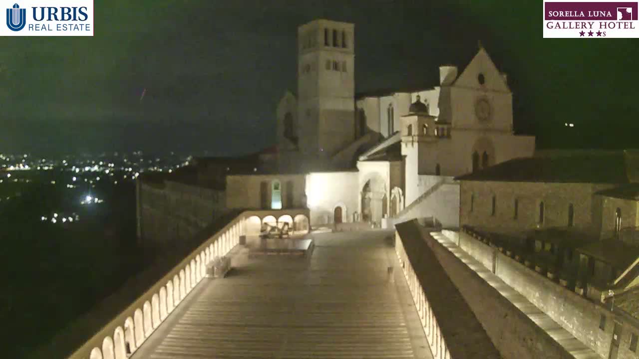 A large, illuminated stone building complex, possibly a church or monastery, is situated on a hill at night, overlooking a city with a dark, clear sky.