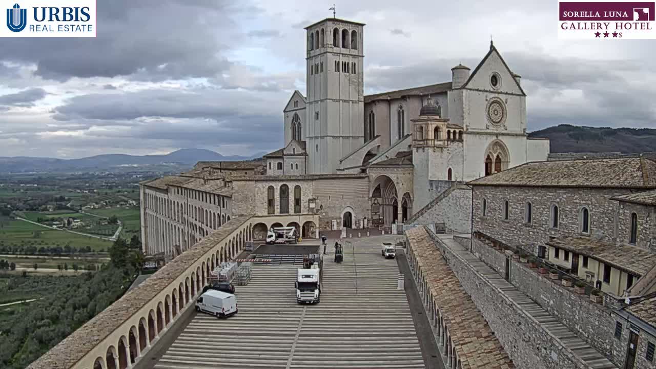 A large, light-colored basilica and adjacent buildings are situated on a hill overlooking a valley under a cloudy sky, with several vehicles parked in a paved area between the structures.