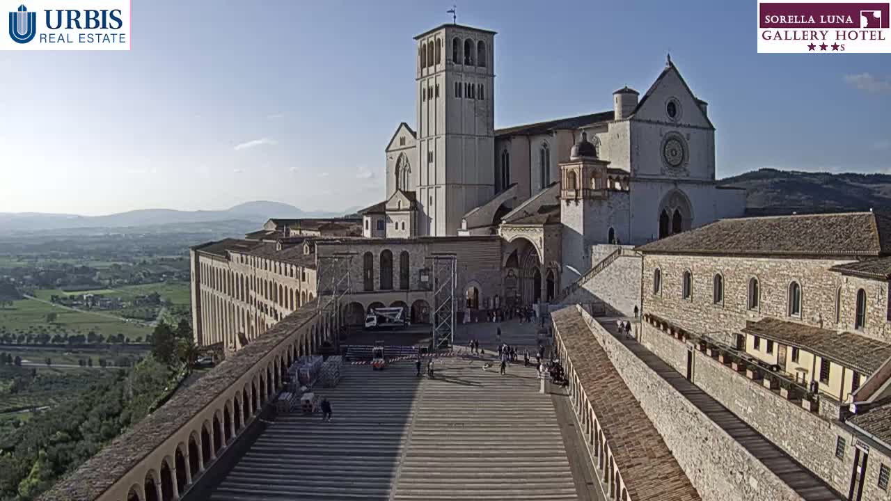 A large stone basilica and adjoining buildings are seen from above on a sunny day, overlooking a valley.