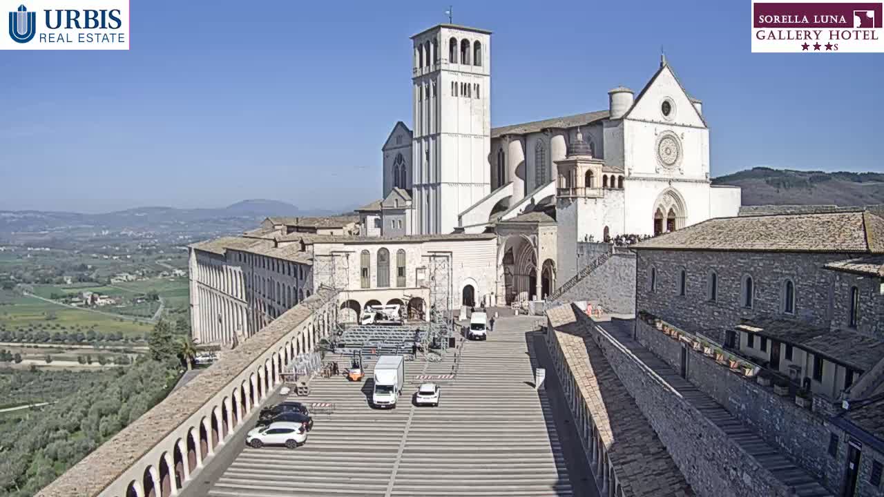 A large, light-colored basilica stands on a hill overlooking a valley under a clear blue sky, with a paved plaza in front and several vehicles parked.
