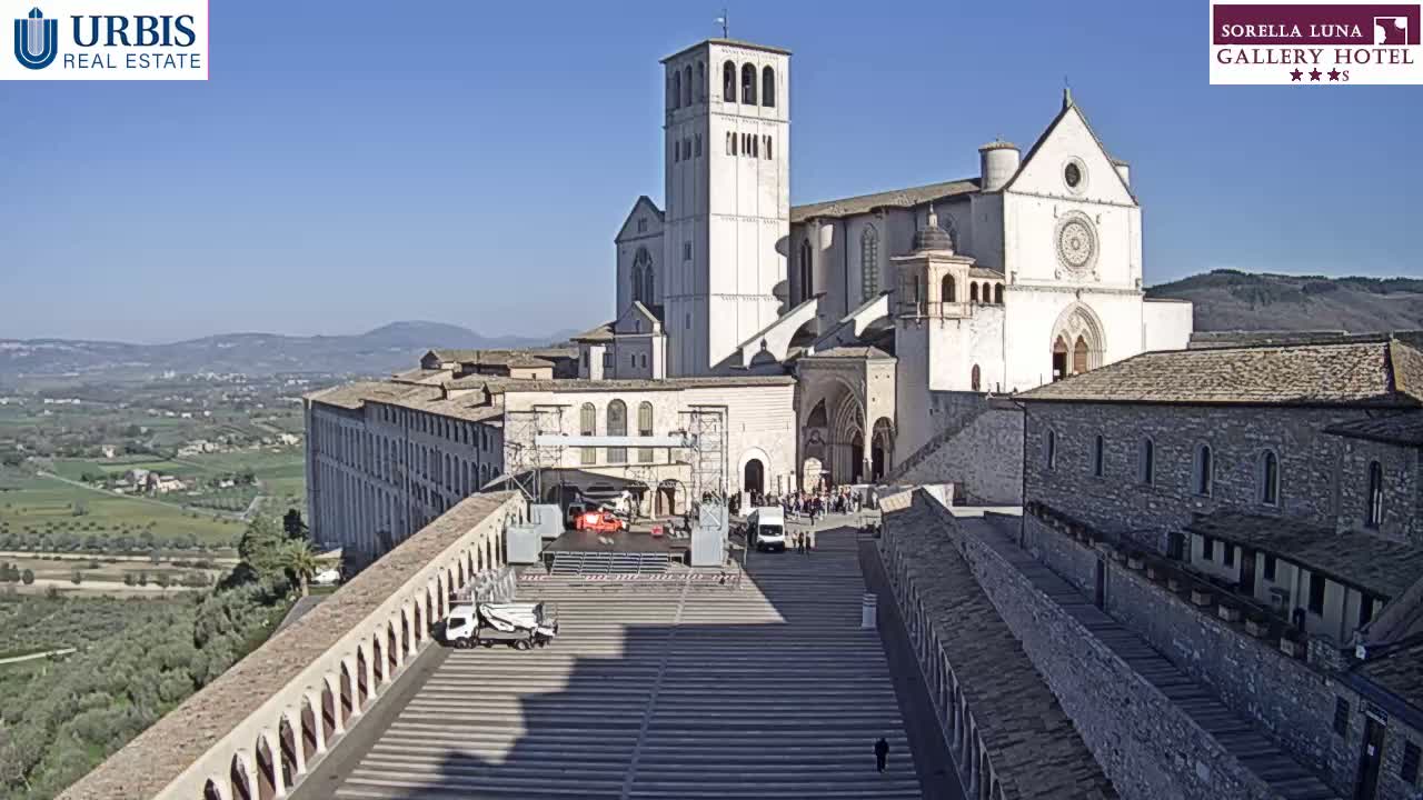 A large, white basilica sits atop a hill overlooking a valley on a sunny day, with a plaza in the foreground featuring a stage and several vehicles.
