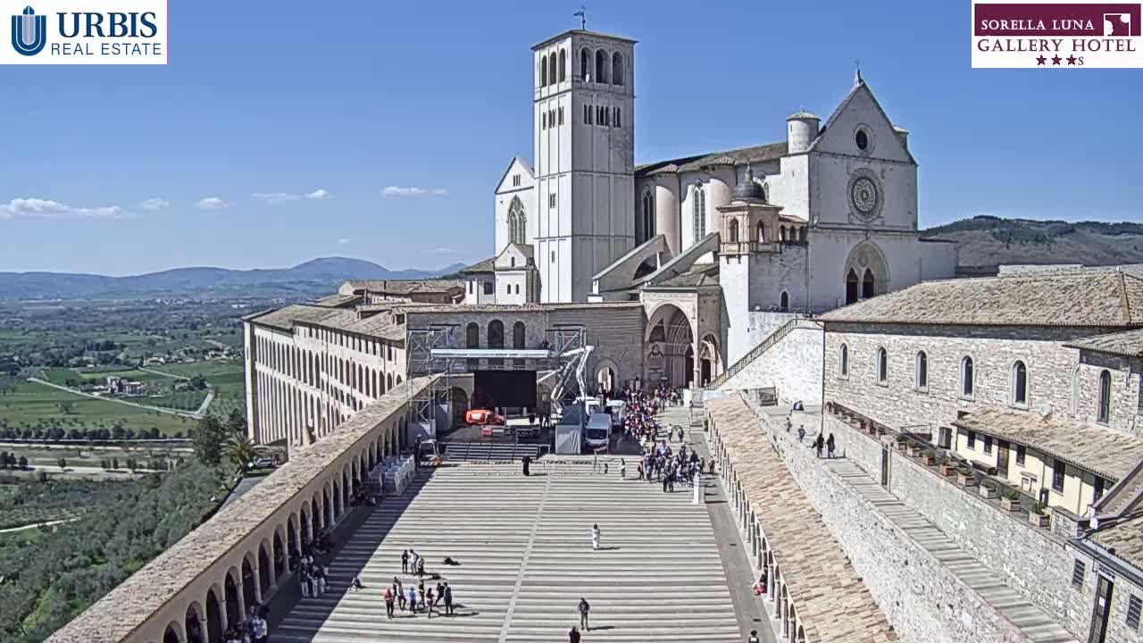 A large stone basilica and adjoining buildings are viewed from above on a sunny day, with a plaza below containing a stage and a number of people.