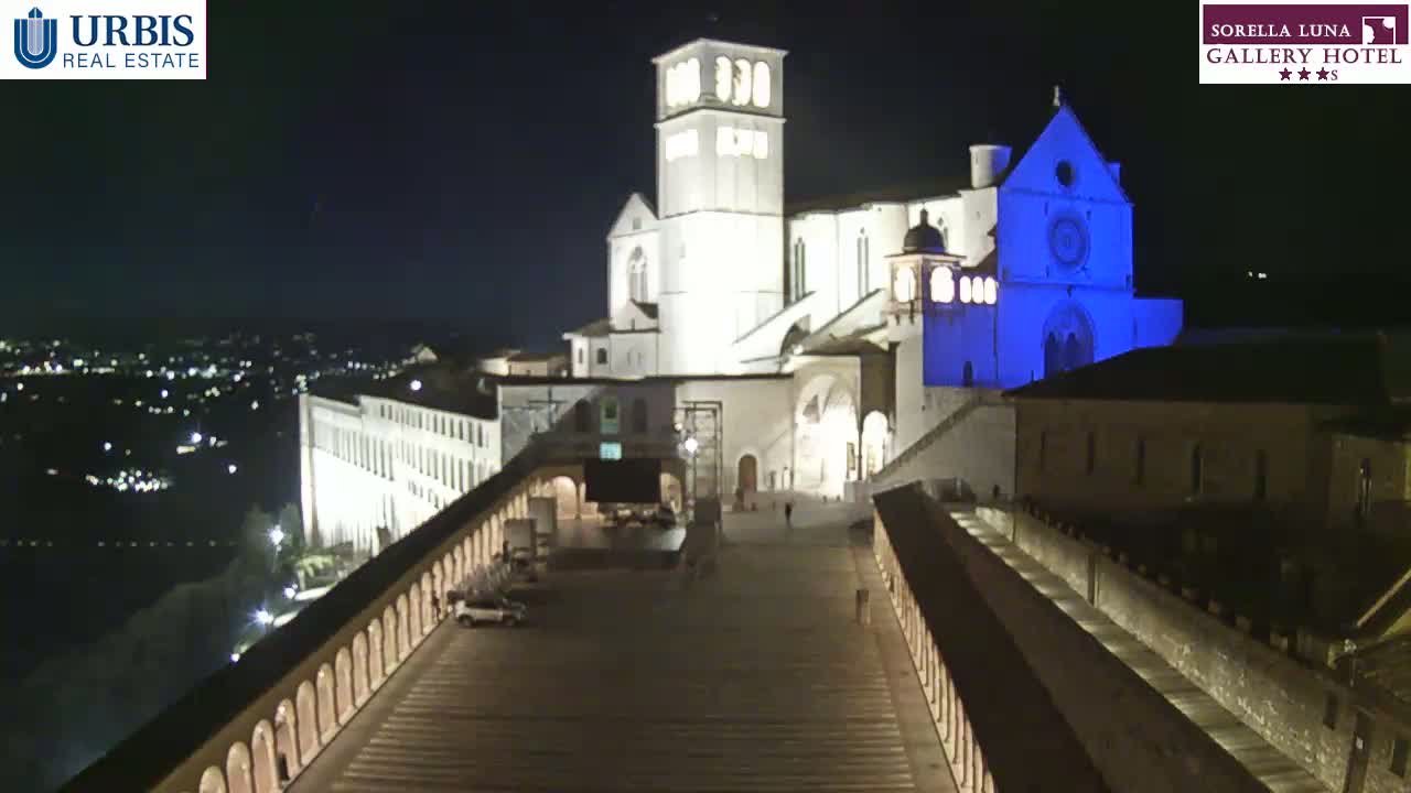 A large, illuminated basilica stands at night on a hill overlooking a city, with a long, paved walkway leading down from it.