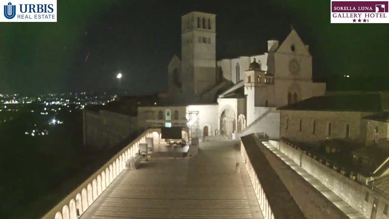 A nighttime view of a large stone building complex, possibly a church or monastery, overlooking a city illuminated by lights, under a clear, moonlit sky.