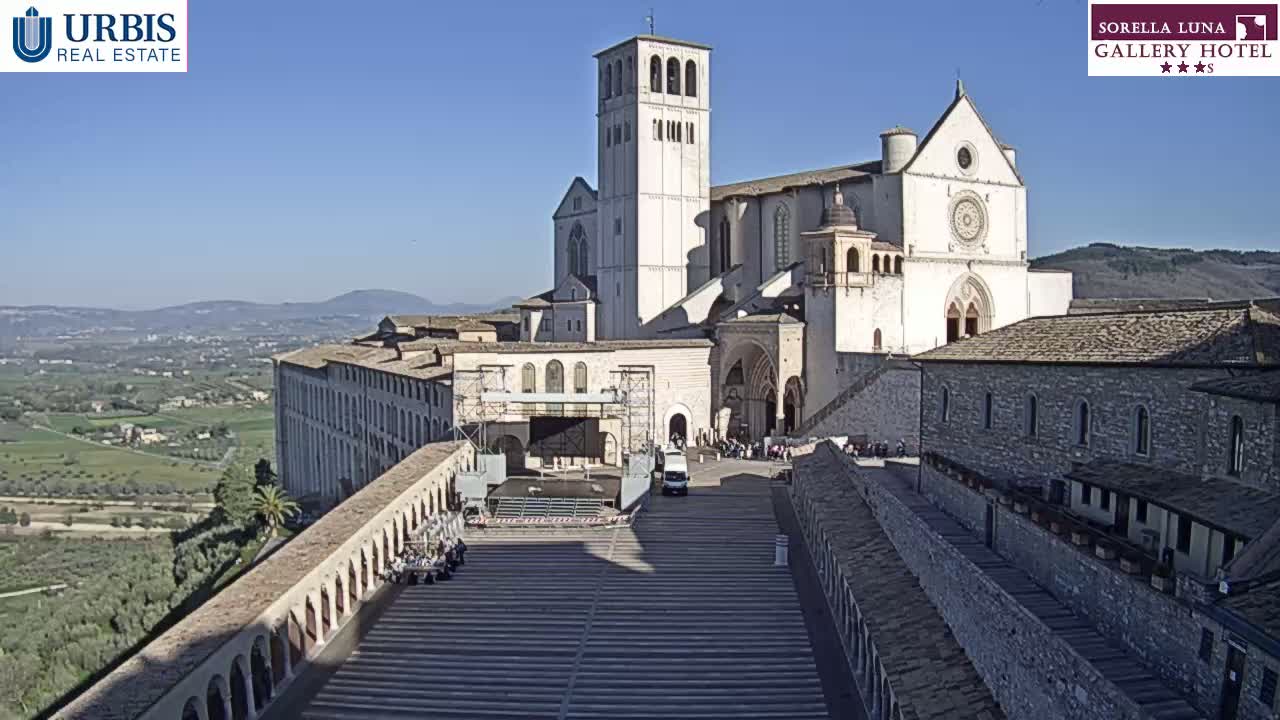 A large stone basilica overlooks a paved plaza and a valley on a sunny day.